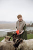 A woman in a green dress and beige shawl stands outdoors holding a red bucket, surrounded by sheep in a rural landscape with hills and a house in the background.