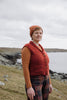 A woman standing outdoors on a grassy coastal hill under an overcast sky, wearing a rust-orange cable-knit vest over a rust-orange long-sleeve top, with plaid trousers.