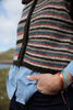 A close-up of a person wearing a striped knit cardigan with dark buttons over a light blue button-up shirt and dark jeans, with a blurred outdoor background.