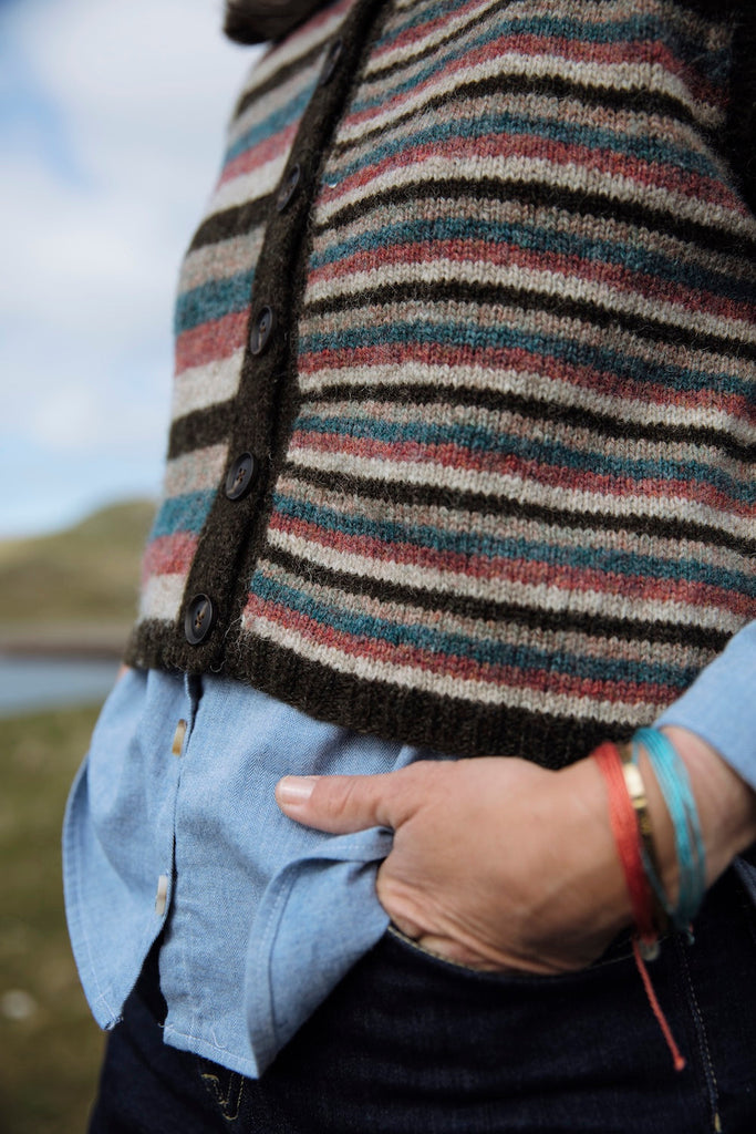 A close-up of a person wearing a striped knit cardigan with dark buttons over a light blue button-up shirt and dark jeans, with a blurred outdoor background.