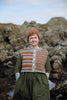 A woman standing outdoors on a rocky shoreline wearing a handmade striped knit vest over a green dress and an orange beanie.