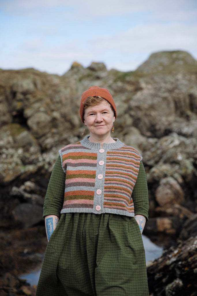 A woman standing outdoors on a rocky shoreline wearing a handmade striped knit vest over a green dress and an orange beanie.