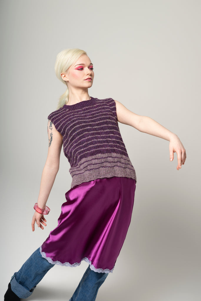 Person wearing a sleeveless purple top and skirt with blue jeans on a gray background