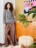 Woman in a patterned top and brown skirt standing in a room with wooden furniture and plants.