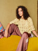 Woman sitting on a yellow couch wearing a beige crochet top and brown pants.
