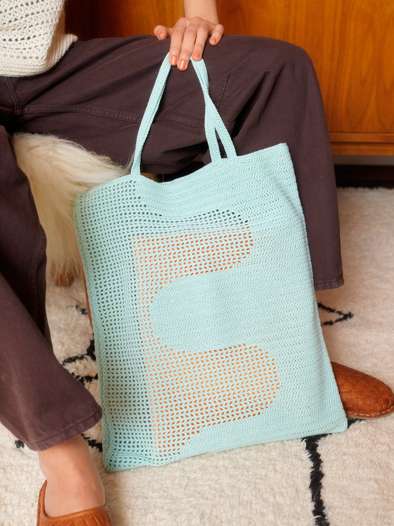 Person holding a light blue mesh tote bag on a patterned rug.