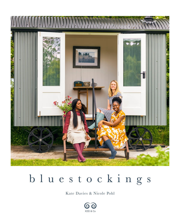 The image features the cover of the book "Bluestockings" by Kate Davies and Nicole Pohl. The cover shows two women seated outside a cozy, rustic-looking structure that resembles a shepherd's hut.